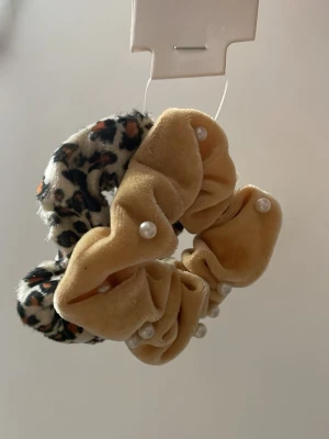 Scrunchies med pärlor & leopardmönster - Två scrunchies i mjuk sammet, en i beige med små pärlor och en med leopardmönster i svart, beige och orange. Perfekta för att ge din frisyr en trendig och lekfull touch.