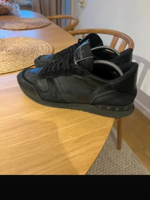 Svarta Valentino rockrunners Garavani sneakers - Snygga helsvarta sneakers från Valentino Garavani med coola nitar på sulan. Skorna är gjorda i en mix av mocka och läder, har låga skaft och klassisk snörning. Perfekta för dig som gillar stilrena och exklusiva detaljer. skosnörena är lite slitna