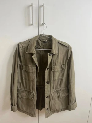 Grön overshirt med knappar - Stilren grön overshirt med fyra fickor framtill och knappar längs hela framsidan. Jackan har klassisk krage, långa ärmar och justerbar midja med snörning. Perfekt för lager på lager och enkel att matcha.