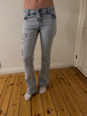 Ljusblå jeans med fjärilsbrodyr - Säljer ett par ljusblå jeans med raka ben och snygg fjärilsbrodyr på sidan av benet och bakfickan. Jeansen har klassisk femficksmodell, normal midja och är tillverkade i denim. Perfekta för dig som vill ha en unik och trendig look.