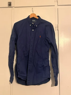 Marinblå skjorta från Polo Ralph Lauren - Säljer en klassisk mörkblå skjorta från Polo Ralph Lauren med orange broderad logga på bröstet. Skjortan har knappar hela vägen och en button-down krage. Nypris 1799 kr.