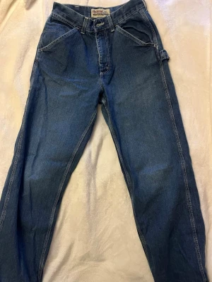 Blå baggy jeans från Crocker - Säljer ett par blå baggy jeans från Crocker med raka ben, stora fickor bak och coola detaljer på sidorna. Klassisk femficksmodell i denim med hällor för bälte och robust känsla. 