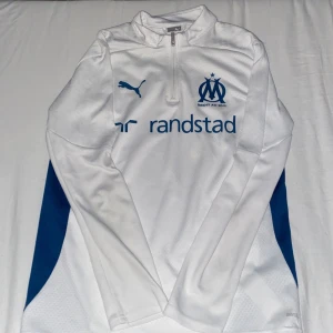 Vit långärmad Marseille Puma tröja - Säljer en vit långärmad fotbollströja från Puma med Olympique de Marseille-logga och blå detaljer på sidorna. Tröjan har halv dragkedja fram, tryckt Puma-logga och sponsortryck. Materialet är lätt och ventilerande polyester, perfekt för träning.