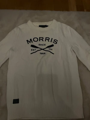 Vit stickad tröja från Morris - Vit stickad tröja från Morris med svart tryck framtill där det står 'Morris Since 1968' och två korsade åror. Tröjan har rund halsringning och ribbade muddar vid ärmslut och nederkant. Perfekt för en stilren och sportig look.