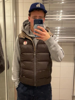 Brun dunväst från Colmar Originals - Snygg brun dunväst från Colmar Originals med beige detaljer och  luva. Västen har dragkedja framtill, två fickor med dragkedja och Colmar-logga på bröstet. Märket på bröstet blev utslitet pga tvätt, annars inga defekter eller hål. Passar XS/S. väldigt snygg och stilren. Skickas i kartong. 