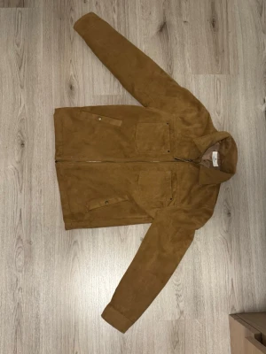 Brun overshirt i mocka - Snygg brun overshirt i mockaimitation med två stora bröstfickor och knappdetaljer vid ärmsluten. Jackan har rak passform, klassisk krage och dragkedja framtill. Perfekt lager på lager-plagg för en trendig look.