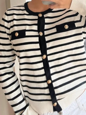 Randig kofta med guldknappar - Snygg vit kofta med svarta horisontella ränder och dekorativa guldknappar framtill. Två fejkade bröstfickor med svarta detaljer. Långärmad och rund halsringning. Perfekt för en trendig och stilren look.