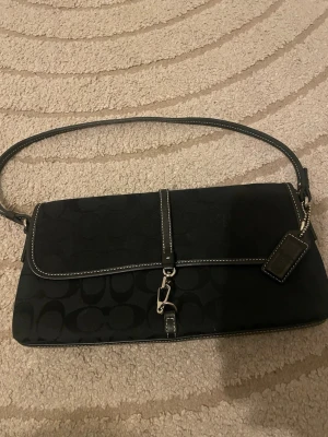 Svart axelväska från Coach. ÄKTA - Snygg svart axelväska från Coach med klassiskt monogrammönster, silverfärgat spänne och detaljer i läder. Väskan har en kort axelrem och en tag med Coach-logga. Perfekt för dig som gillar stilrena accessoarer. Äkta