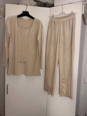Beige loungewear-set i mjuk velour - Mysigt loungewear-set i beige velour. Setet består av en långärmad öppen cardigan, ett linne med rund hals och ett par byxor med resår i midjan. Perfekt för chill dagar hemma och riktigt skönt material. S/M oanvänd 