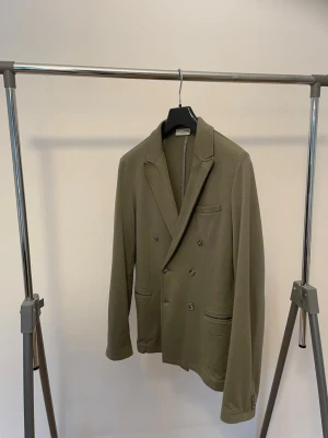 Olivgrön dubbelknäppt kavaj - Stilren Paolo Pecora olivgrön kavaj med dubbelknäppning och klassiska slag. Kavajen har tre framfickor och dekorativa knappar vid ärmsluten. Tillverkad i ett mjukt tyg som ger en avslappnad men ändå dressad look. Pris kan diskuteras vid snabb affär.