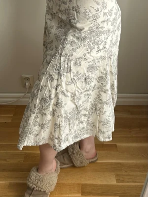 Vit långkjol med blommigt mönster - Säljer en vit långkjol med grått blommigt mönster. Kjolen har en lätt utsvängd form och är gjord i ett mjukt, tunt material som känns skönt mot huden. Perfekt för dig som gillar romantiska och feminina plagg. En gul fläck, men inget jag har tänkt på