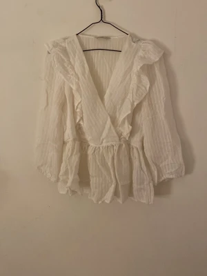 Vit omlottblus med volanger - Söt vit omlottblus med volangdetaljer och v-ringning som går ganska långt ner. Blusen har kortare ärmar och är tillverkad i ett lätt, tunt tyg med diskreta ränder. (Blusen är lite skrynklig i bilden så det syns inte så väl)