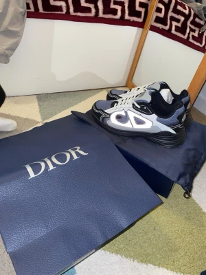 D**e B22 sneakers svart/vit - Säljer ett par Dior B22 sneakers i svart, vitt och grått med chunky sula och meshdetaljer. Skorna har en modern, sportig design med tydliga Dior-loggor på sidan och bak. Kombination av läder och mesh ger både stil och komfort. Perfekta för dig som gillar exklusiva sneakers.