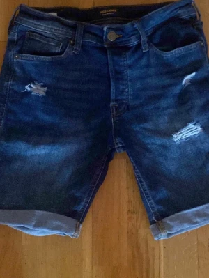 Blå jeansshorts från Jack & Jones - Snygga blå jeansshorts från Jack & Jones med slitna detaljer och upprullade benslut. Klassisk femficksmodell med knapp och dragkedja. Perfekta för en avslappnad stil under varma dagar.