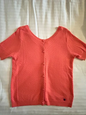 Coralfärgad stickad T-shirt - Kortärmad kofta i korallrött med rund halsringning och knappar framtill. Stickad struktur med diskret mönster och ribbade kanter vid ärmslut och nederkant.