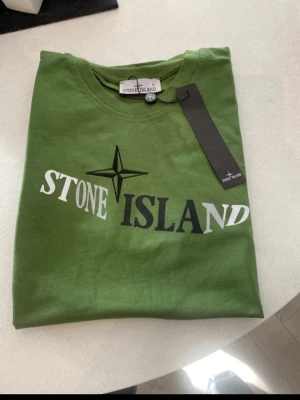 Grön Stone Island t-shirt med logga - Snygg grön t-shirt från Stone Island med stor logga och text på bröstet i vitt och svart. Klassisk rund halsringning och rak passform. Tillverkad i mjuk bomull som känns skön mot huden. Perfekt för dig som gillar streetwear och vill sticka ut.