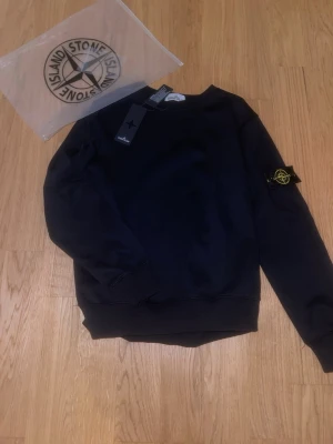 Svart sweatshirt från Stone Island - Svart sweatshirt från Stone Island med klassisk rund hals och ribbade muddar. Ikonisk logotyp-patch på vänster ärm. Tröjan är i mjukt material och har en stilren, enkel design som passar perfekt till streetwear-stilen.