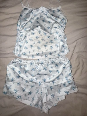 Vit blommig pyjamas-set  - Sött pyjamas-set med blått blommönster. Setet består av ett linne med tunna axelband och spetsdetaljer samt matchande shorts med resår i midjan. Perfekt för mysiga kvällar hemma.