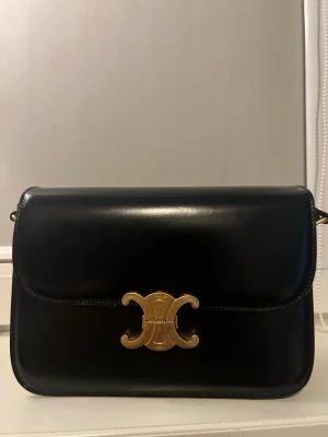 Svart Triomphe handväska från Celine - Snygg svart handväska i slätt läder från Celine med ikoniskt guldigt Triomphe-spänne framtill. Klassisk rektangulär form och minimalistisk design som ger en lyxig känsla. Perfekt för dig som gillar stilrena accessoarer med exklusiva detaljer.
