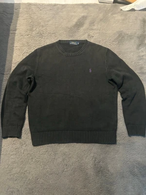 Mörkgrå stickad tröja Polo Ralph Lauren - Snygg mörkgrå stickad tröja från Polo Ralph Lauren med lila broderad logga på bröstet. Tröjan har rund halsringning och ribbstickade muddar vid ärmslut och nederkant. Perfekt för en stilren och avslappnad look.