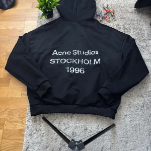 Svart hoodie Acne Studios Stockholm - Svart hoodie från Acne Studios med stor huva och tryck i vitt på ryggen där det står 'Acne Studios STOCKHOLM 1996'. Klassisk oversized passform med ribbade muddar vid ärmslut och nederkant. Perfekt för en clean och stilren look.