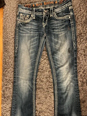 Rock Revival  bootcut jeans - Snygga blå bootcut jeans från Rock Revival med slitna detaljer och fina stenar. De är ett par äkta jeans som är köpta second hand. De är i storlek 26. Och dom är i bra skick❤️