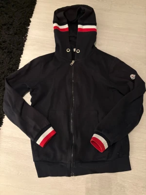 Marinblå hoodie med dragkedja - Snygg marinblå hoodie från Moncler med dragkedja framtill och huva. Röd och vit rand på huvan samt vid ärmsluten ger en sportig vibe. Moncler-logga på ena ärmen. Tillverkad i mjukt bomullsmaterial, perfekt för chill dagar. Priset är inte hugget i sten vid snabb affär! 