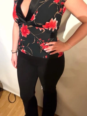 Svart blommig omlottopp med knyt - Snygg svart omlottopp med röda blommor och korta ärmar. Toppen har ett dekorativt knytband i midjan och djup v-ringning framtill. Materialet känns mjukt och stretchigt, perfekt för att skapa en feminin siluett. Passar dig som vill sticka ut med ett färgstarkt mönster.