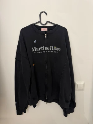 Svart zip sweatshirt från Martine Rose - Cool svart hoodie från Martine Rose med dragkedja framtill och tryckt logga på bröstet. Oversized passform och ribbade muddar vid ärmslut och nederkant. Tillverkad i mjukt bomullsmaterial, perfekt för en avslappnad streetwear-look.