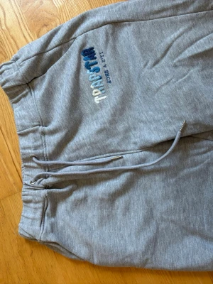 Gråa mjukisbyxor från Trendstyle - Gråa mjukisbyxor från Trendstyle med broderad logga i blått och vitt på ena benet. Byxorna har dragsko i midjan och resår för extra komfort. Perfekta för chill dagar och har en klassisk, avslappnad passform.