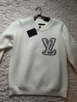 Lv tröja - Snygg vit tröja från Louis Vuitton med stor grå LV-logga på bröstet. Tröjan har rund halsringning, långa ärmar och ribbade muddar. Perfekt för dig som gillar exklusiva märken och stilren design.