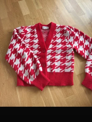 Röd och vit kofta från Monki i XS - Säljer en röd och vit kofta från Monki med stort hundtandsmönster. Koftan har två knappar framtill, ribbade muddar och en croppad passform. Perfekt för dig som gillar statement-plagg och vill sticka ut lite extra.