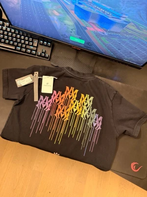 Svart AMIRI t-shirt med färgglatt tryck - Svart t-shirt från AMIRI med coolt flerfärgat drip-tryck på ryggen och mindre tryck framtill. Klassisk passform med rund hals och korta ärmar. Perfekt statement-plagg för dig som gillar streetwear och unika detaljer.