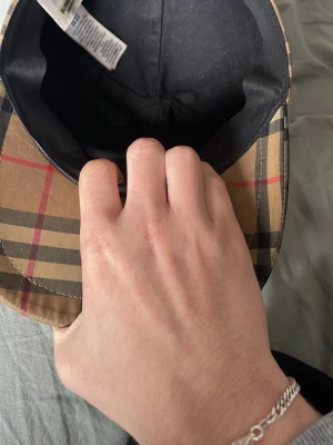 Burberry beige rutig bucket hat - Säljer en klassisk bucket hat från Burberry i beige med det ikoniska rutmönstret i svart, vitt och rött. Hatten är tillverkad i bomull och har svart innerfoder. Perfekt för dig som vill ha en stilren accessoar med lyxig känsla.