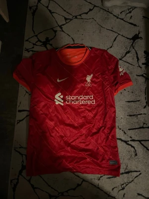 Liverpool matchtröja Nike röd - Säljer en röd Liverpool FC matchtröja från Nike med vita detaljer och klubbmärke på bröstet. Tröjan har korta ärmar, rund hals och sponsorlogga framtill. Materialet är lätt och andas, perfekt för fotbollsträning eller att bära på läktaren.