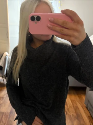 Mörkgrå stickad polotröja vero moda - En snygg mörkgrå stickad polotröja med lång ärm och hög krage. Tröjan har en avslappnad passform och är perfekt för att hålla sig varm under kyliga dagar. Materialet ser ut att vara mjukt och bekvämt, idealiskt för lager-på-lager.