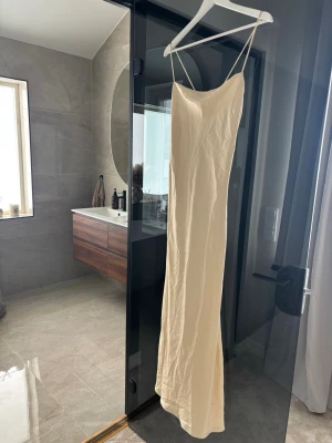 Lång champagnefärgad satin klänning -  elegant långklänning i champagnefärg med tunna axelband och minimalistisk design. Klänningen har en smal passform och är tillverkad i ett mjukt, glansigt material som ger en lyxig känsla. 