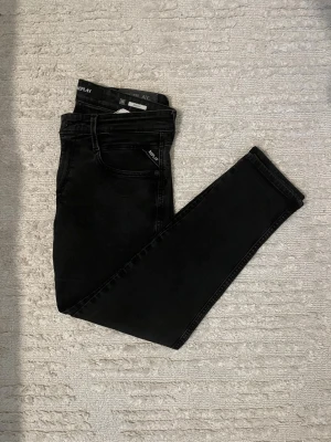 Svarta slim fit jeans från Replay - Säljer ett par svarta slim fit jeans från Replay med klassisk femficksmodell och diskret Replay-logga vid fickan. Jeansen har en snygg tvättad look och är tillverkade i stretchigt denimtyg för extra komfort.