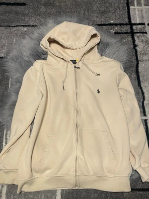 Beige hoodie från Polo Ralph Lauren - Snygg beige hoodie från Polo Ralph Lauren med dragkedja, två fickor framtill och klassisk svart broderad logga på bröstet. Hoodien har justerbar huva med snören och ribbade muddar vid ärmar och nederkant. Perfekt för en chill och stilren look. Den är använd 3 gånger säljs eftersom använder den inte längre och priset går att diskuteras
