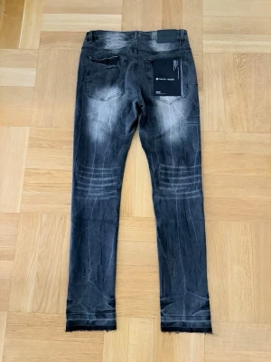 Svarta skinny jeans från Purple Brand - Säljer ett par svarta skinny jeans från Purple Brand med slitna detaljer och tvättad look. Jeansen har råa kanter vid bensluten, fem fickor och silverfärgade nitar. Materialet är denim med stretch för extra komfort. Perfekta för en trendig streetwear-stil.