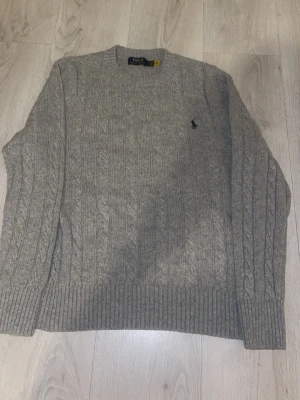 Grå kabelstickad tröja Polo Ralph Lauren - Kabelstickad grå tröja från Polo Ralph Lauren med klassisk rund halsringning och broderad marinblå logga på bröstet. Tröjan har ribbade muddar vid ärmslut och nederkant och är tillverkad i mjukt garn för en skön känsla.