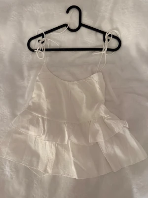 Vit volang topp med tunna band - Supersöt vit linnetopp med tunna axelband som knyts på axlarna och dubbla volanger. Toppen har en luftig och flowy känsla, perfekt för varma dagar. Passar till jeans, kjol eller shorts för en fräsch sommarlook.