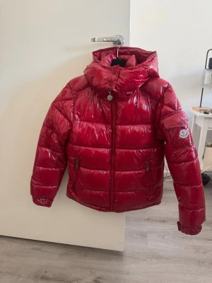 Röd glansig dunjacka från Moncler - Snygg röd dunjacka från Moncler med glansig finish och stor huva. Jackan har två dragkedjefickor fram och klassisk Moncler-logga på ärmen. Perfekt för kalla vinterdagar och ger en riktigt trendig look.