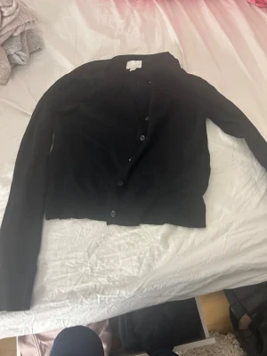 Svart kofta från H&M med knappar - En klassisk svart kofta från H&M med långa ärmar och knäppning framtill. Koftan är ribbad vid ärmslut och nederkant, vilket ger en snygg passform. Perfekt att slänga över en topp eller klänning för en enkel och stilren look.