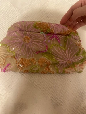 Blommig broderad kuvertväska - Kuvertväska med broderade blommor i lila, orange och grönt på beige mesh. Väskan har paljettdetaljer och en mjuk, rundad form. Perfekt för dig som gillar färg och unika accessoarer.