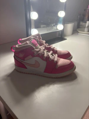 Nike Air Jordan 1 Mid rosa/vit - Snygga Nike Air Jordan 1 Mid sneakers i rosa och vitt med klassisk swoosh på sidan. Skorna har rosa detaljer, perforerad tåbox och vita skosnören. Tillverkade i läder och textil för en bekväm och stilren look. Perfekta för dig som vill sticka ut med färg.