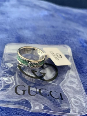 Gucci silverring med grön detalj - Snygg ring från Gucci i äkta silver (Ag 925) med en grön marmorerad detalj runt hela ringen. Ringen har en klassisk, rundad form och Guccis ikoniska logga framtill. Perfekt för dig som vill ha något unikt och stilrent från ett lyxigt märke. Storlek 16.
