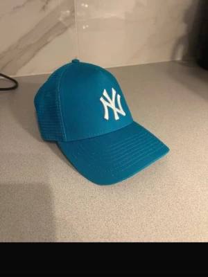 Blå New Era NY keps - Snygg blå keps från New Era med klassiskt New York Yankees-logga broderad i vitt framtill. Kepsen har mesh-paneler på sidorna för extra ventilation och böjd skärm. Perfekt för dig som vill ha en sportig och trendig look.