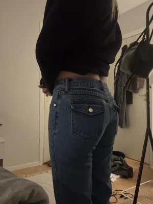 Blå raka jeans med hög midja - Säljer ett par klassiska blå jeans med hög midja och raka ben. Jeansen har fem fickor och silverfärgade knappar på bakfickorna. Perfekta till vardags och passar till det mesta. Materialet är denim i bomull.