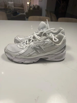 Vita New Balance sneakers - Snygga vita sneakers från New Balance med mesh- och syntetmaterial. Skorna har en sportig design med silvriga detaljer och klassisk N-logga på sidan. Perfekta för dig som gillar en clean och trendig look.